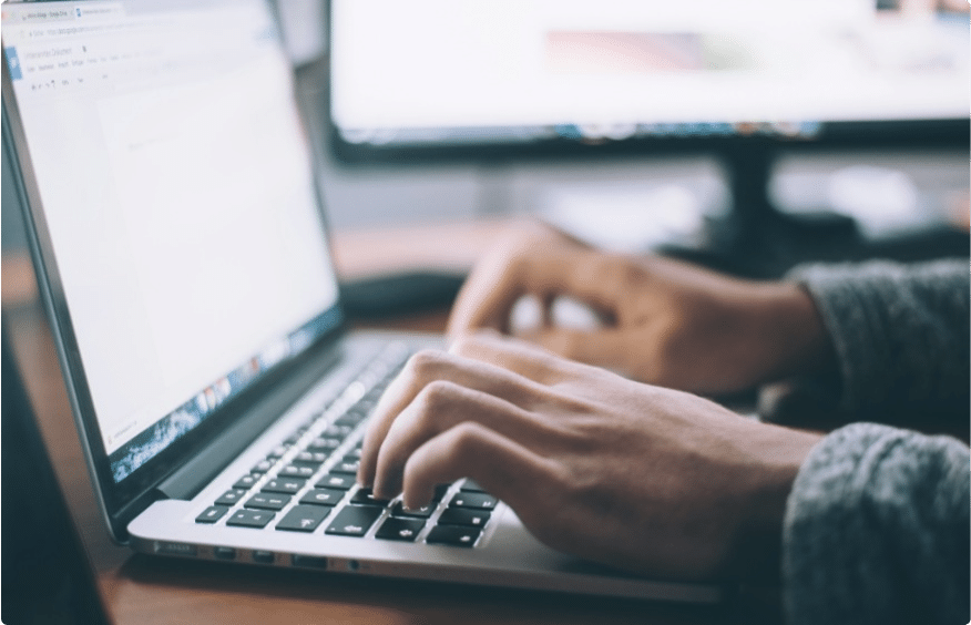 A person is typing on a laptop keyboard, with a computer monitor visible in the background. The laptop screen displays an open document, and both screens give off a soft glow. The person's sleeves are long, suggesting a casual or cozy setting.